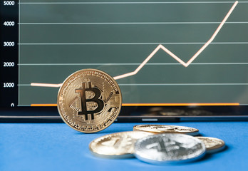 Few gold coins of bitcoins lie on the table in front of and next to the tablet on which charts of Bitcoin's cost growth are visible. One silver bitcoin stands side by side on the edge. 