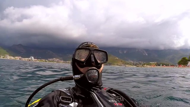 Male diver in scuba diving equipment floating on waves on water surface and waving.  Scenic island on background
