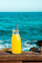Glass bottle with citrus fruit juice orange with straw on wooden rail. Turquoise sea and blue sky in the background. Tropical vacation summer clean eating concept