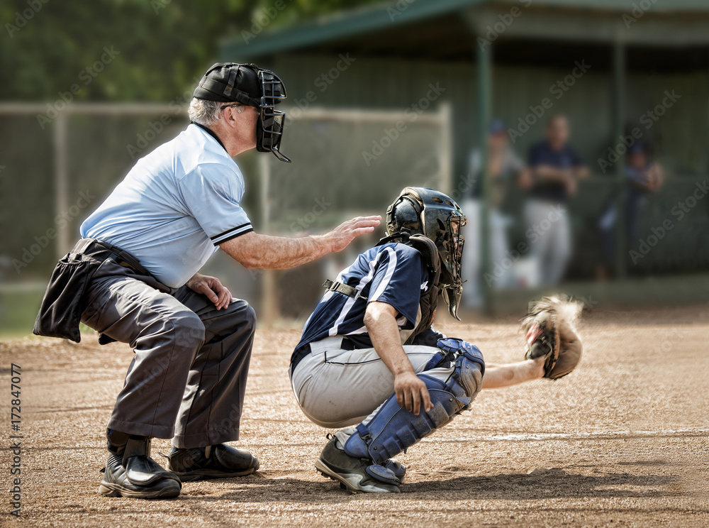 Wall mural Umpire and catcher, just as ball hits catcher's mitt, dugout and players in background