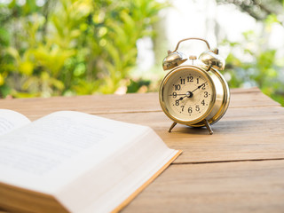 text book with vintage alarm clock on the wood table. The background is green from tree and light bokeh