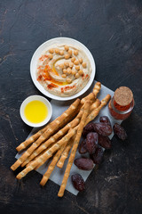Homemade hummus with grissini and date fruits on a dark brown stone surface, flat-lay, vertical shot