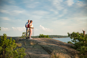 Active senior couple hiking on the top of rock, exploring. Mature man and woman Happily smiling. Nordic walking, trekking. Scenic view of gulf and sea. Healthy lifestyle. Finland.