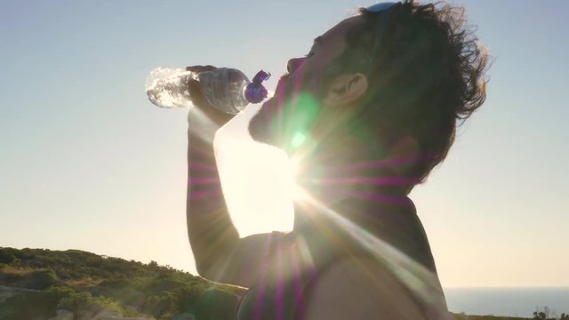 Runner arrives on top of a cliff on an island in front of the ocean and drinks water from a plastic bottle