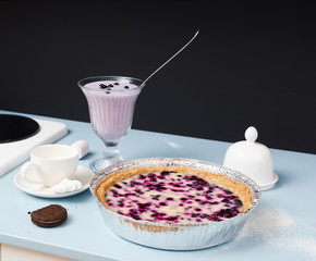 Homemade blueberry pie with cream filling served with blueberry smoothie, chocolate cookie, marshmellows and milk on pale blue table with black background