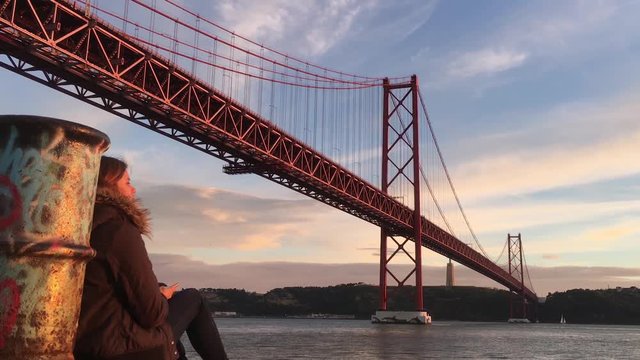 Girl Enjoying Time Alone Under Famous Bridge of Lisbon. Young woman enjoying beautiful sunset by Tagus river and famous 25 de Abril bridge in Lisbon