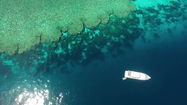 An aerial shot of the great barrier reef. A white small ship can be seen in the middle of the blue ocean.
