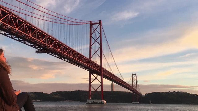 Girl Siting By Tagus River, Zoom Out From 25 Abril Bridge. Young woman enjoying beautiful sunset by Tagus river and famous 25 de Abril bridge in Lisbon