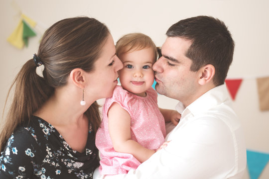 Mother And Father Snuggling With Their Little Girl And Kissing Her