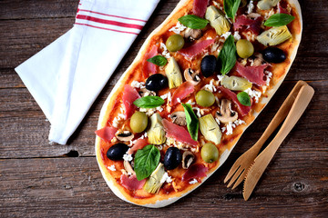 Delicious pizza with artichokes, olives, jamon (hamon), mushrooms, cheese and basil on a wooden background. © chudo2307