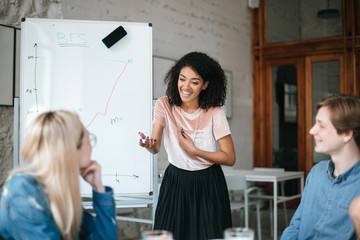 Naklejka premium Portrait of happy African American girl standing near board and discussing new project with colleagues in office . Group of smiling people sitting at the table and listening business presentation