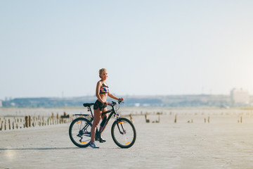 Fototapeta na wymiar A strong blonde woman in a multicolored suit sits on a bicycle in a desert area and looks at the sun. Fitness concept. Blue sky background