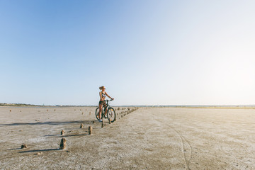 Fototapeta premium A strong blonde woman in a multicolored suit and sunglasses sits on a bicycle in a desert area and looks at the sun. Fitness concept. Blue sky background