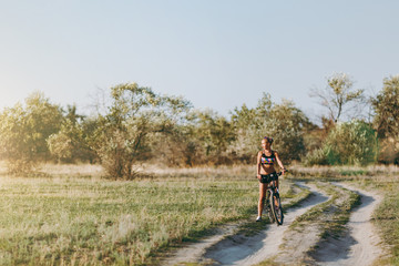 A strong blonde woman in a colorful suit sits on a bicycle in a desert area with trees and green grass and looks at the sun. Fitness concept.