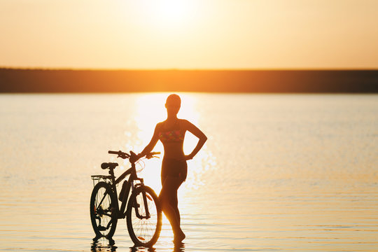 A strong blonde woman in a colorful suit stands near the bicycle in the water at sunset on a warm summer day. Fitness concept. Sky background