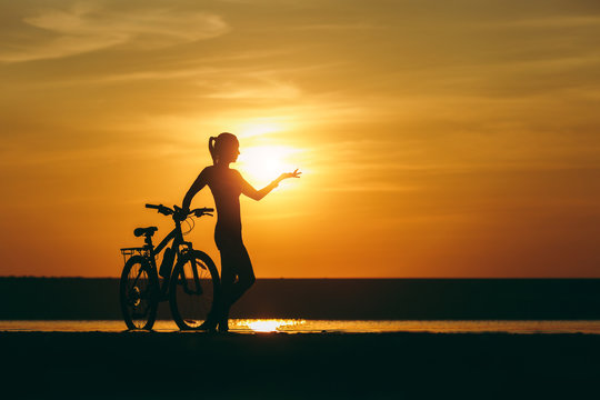 Silhouette of a sporty girl in a suit standing near a bicycle in the water and points her hand to the distance at sunset on a warm summer day. Fitness concept. Sky background