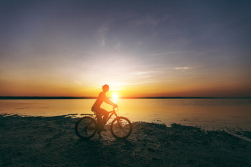 A sporty blonde woman in a colorful suit rides a bike in a desert area near the water on a sunny summer day. Fitness concept. Blue sky background