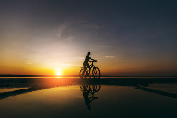 Naklejka na ściany i meble Silhouette of a sporty girl in a suit that sits on a bicycle in the water at sunset on a warm summer day. Fitness concept. Sky background