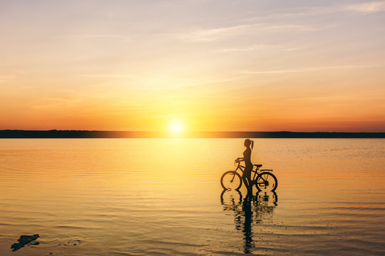 Silhouette of a sporty girl in a suit that sits on a bicycle in the water at sunset on a warm summer day. Fitness concept. Sky background