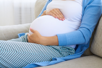 Hands of pregnant woman touch her belly while sitting on sofa
