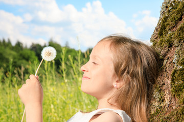 young girl sit next to a tree and smile into the summer sun. she plays with electronic devices, red apple, flowers. sky is blue and the grass is green.