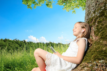 Naklejka na ściany i meble young girl sit next to a tree and smile into the summer sun. she plays with electronic devices, red apple, flowers. sky is blue and the grass is green.