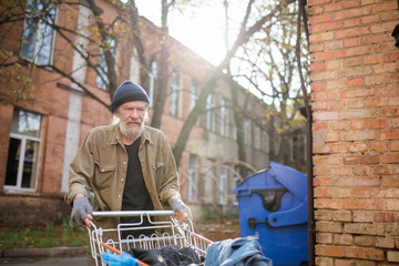 View of homeless old man with cart from supermarket. Old tramp using shopping cart to collect empty...