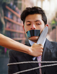 Businessman trapped in a chair with rope, with a black tape in his mouth, afraid of a hand that is...