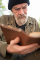 Obraz premium Close up portrait of old beardy man holding book in hands. Wrinkled old male reading a book outdoors.