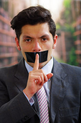 Businessman, with a black tape in his mouth doing a silence sign with a finger in from of his...