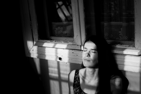 Young woman basking in the rays of the sun in the shadow of the window frames on the porch. Black-and-white photography.