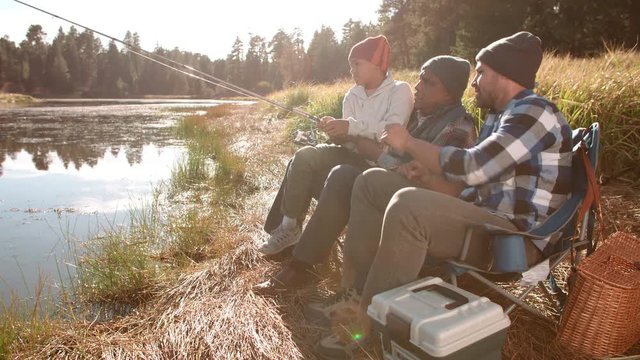 Grandfather and father sit showing boy how to fish by a lake