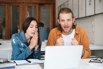 Portrait of joyful man happily looking on laptop while girl sitting near and showing folded hands gesture. Students sitting in classroom and studying together with laptop and books on table