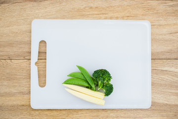 Fresh broccoli, baby corn and sweet peas on white plastic chopping block with wood table background. Copy space