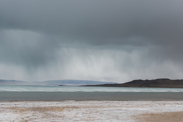panoramic view of lake with snow on coast and storm clouds in sky
