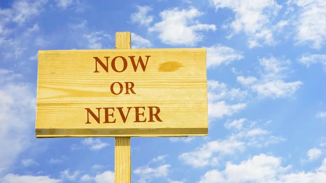 Now or never. Words on a wooden sign against time lapse clouds in the blue sky.