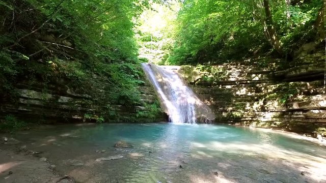 General front view of natural waterfall flowing from rocks into a small lake, surrounded with green trees.
