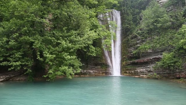 General front view of natural waterfall flowing from rocks into a small lake, surrounded with green trees.