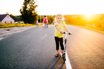 Little beautiful girl with lovely face riding kick scooter outdoor on weekend at sunset. Countryside autumn evening. Kid on road. Russian village lifestyle. Sun rays and shadows. Sincere emotions.