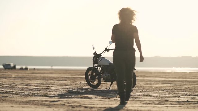 Attractive beautiful woman goes to her old cafe racer motorcycle, sits on it and rides away. Female motorcycle rider. Slow motion