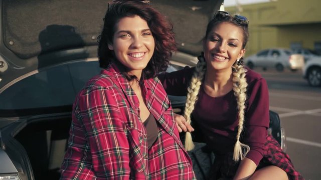 Closeup shot of two young beautiful women sitting in a car trunk in the parking by the shopping mall during sunny day, smiling and looking in the camera. Slowmotion shot