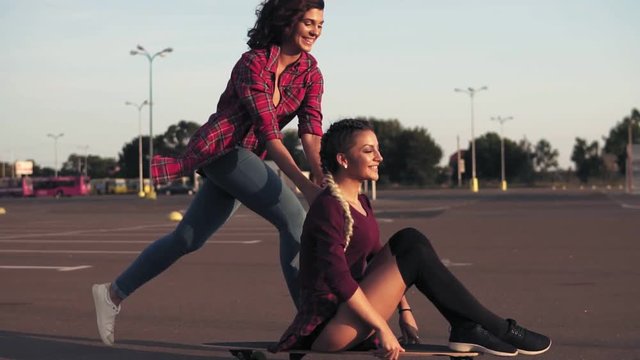 Smiling woman sitting on a longboard while her friend is pushing her behind and running during sunset. Enjoying life. Lens flare. Slowmotion shot
