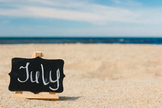 A small wooden malbert with a slate on which chalk is written "august". The concept of summer month, calendar. Beach, golden sand, blue Baltic sea, blue sky.