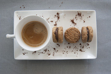top view of ceramic cup with espresso coffee on rectangular white saucer with lady's kisses and chocolate flakes. On gray tablecloth background.