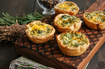 Savory mini quiches (tarts) on a wooden board. Flaky dough pies. Fresh rosemary and dry thyme on a...