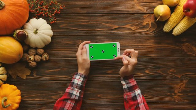 Autumn fruits and vegetables composition on the wooden table. Top view. Woman scrolling, zooming, tapping on the white smartphone with green screen. Horizontal position. Chroma key. Tracking motion