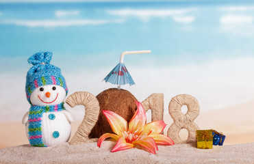 Fototapeta premium New Year inscription 2018, coconut with drinking straw and umbrella instead of number 0, snowman, gifts, flower in sand.