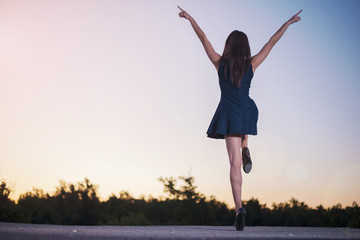 A young slender girl with sexy legs in heels in a miniskirt stands on a sunset background with her hands raised on one leg depicting joyful and vivid emotions.