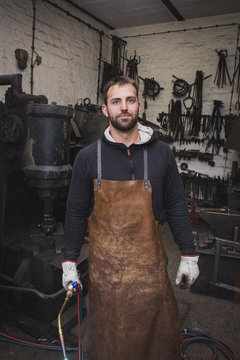 A blacksmith in a leather apron and with a cutting torch is portrayed in his workshop.
