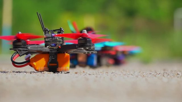 Red and blue racing drone alternately take off from the ground scattering crushed stone in different directions